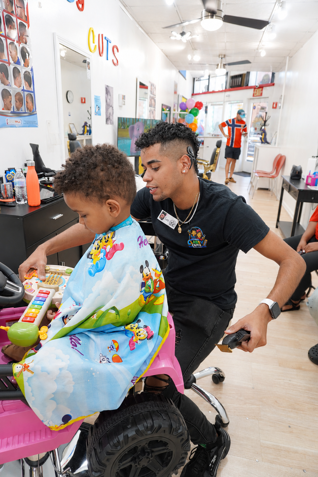 Leo cutting a toddler's hair in a pink car chair with toys